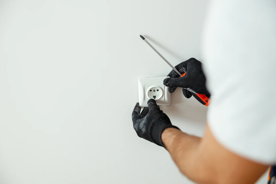 Un électricien en vêtements de travail et ceinture porte-outils en cuir transporte un rouleau de câble électrique blanc dans un environnement de chantier aux murs beiges